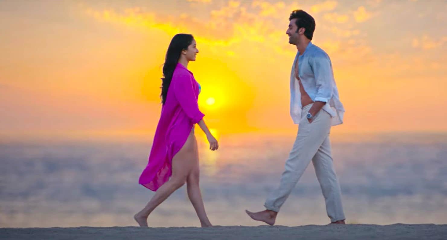 Ranbir Kapoor wearing white shirt and jeans along with Shraddha Kapoor wearing a bikni is walking down the beach during sunset. Still is from the youtube music video  of Tere Pyaar Mein Lyrics